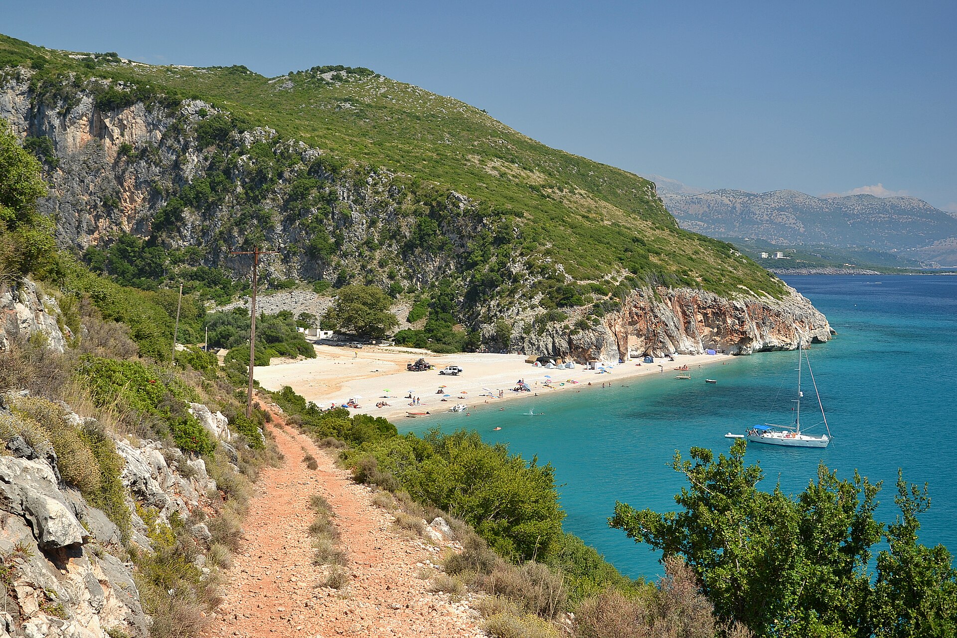 A path above Gjipe Beach with a white cove, turquoise water and a sailboat