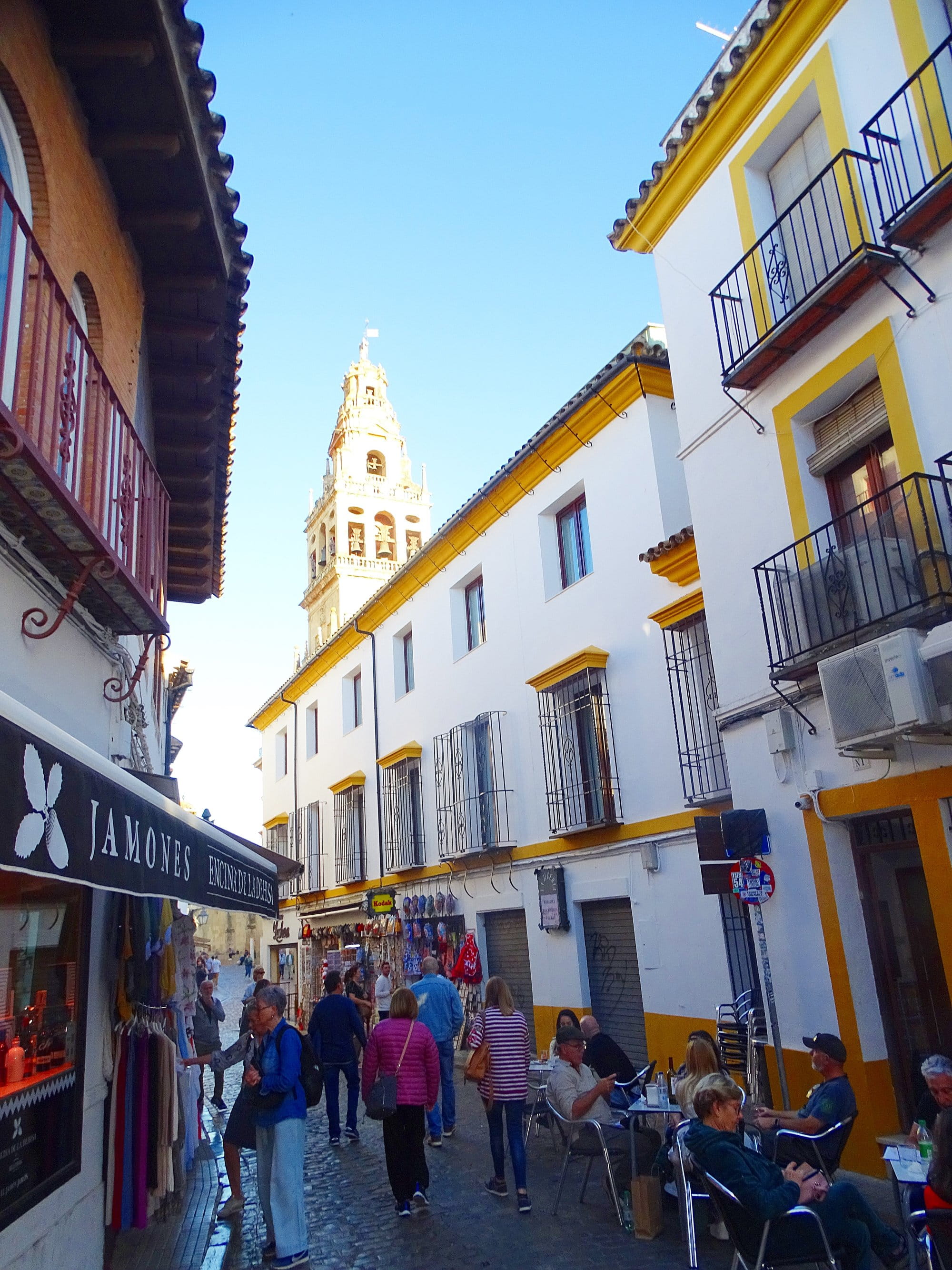 A narrow street in Cordoba's Juderia with white and ochre houses, a shop awning and the Mosque-Cathedral bell tower at the end