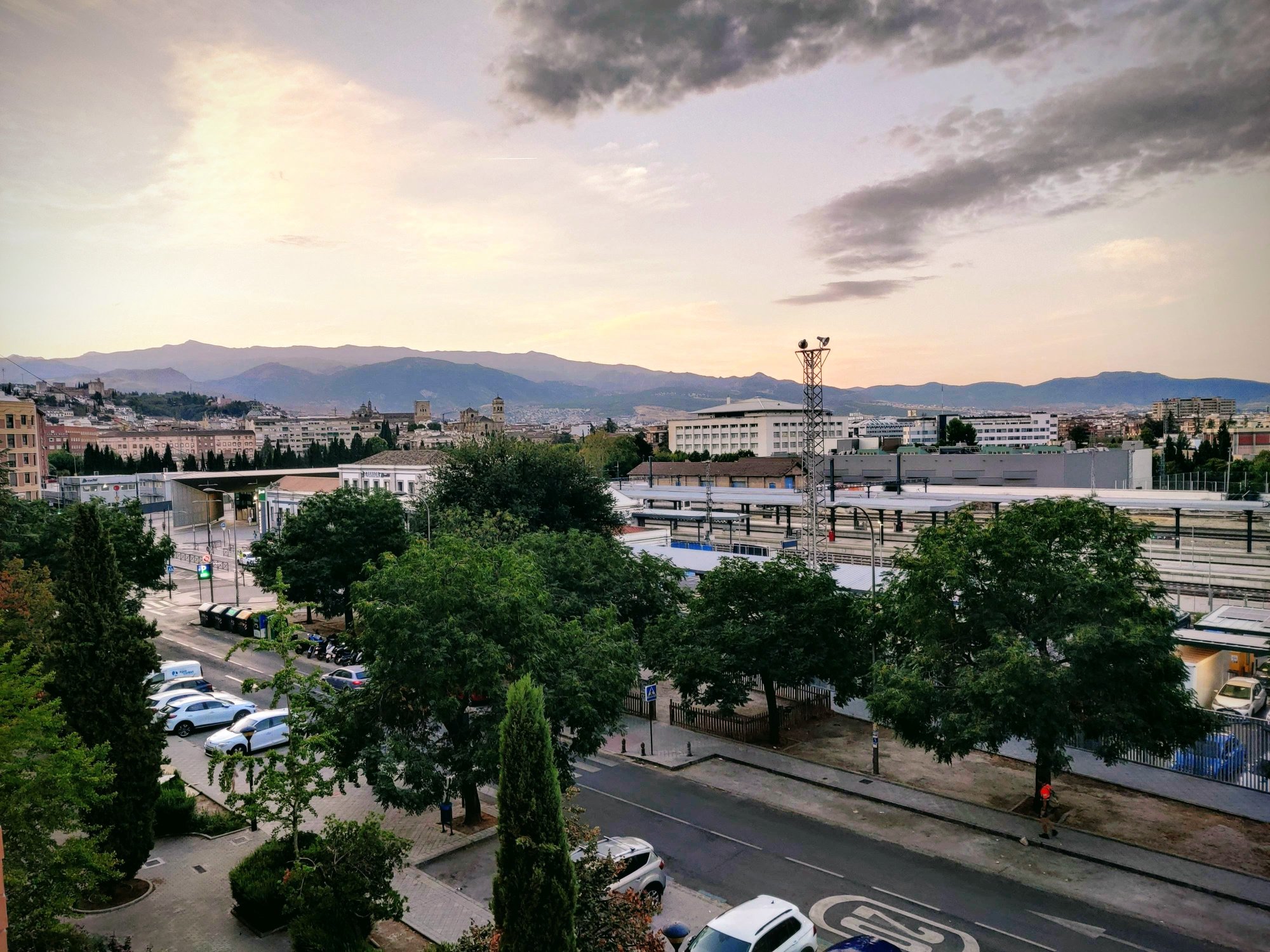 Granada railway station forecourt at dawn with parked cars, trees and the mountains behind the city