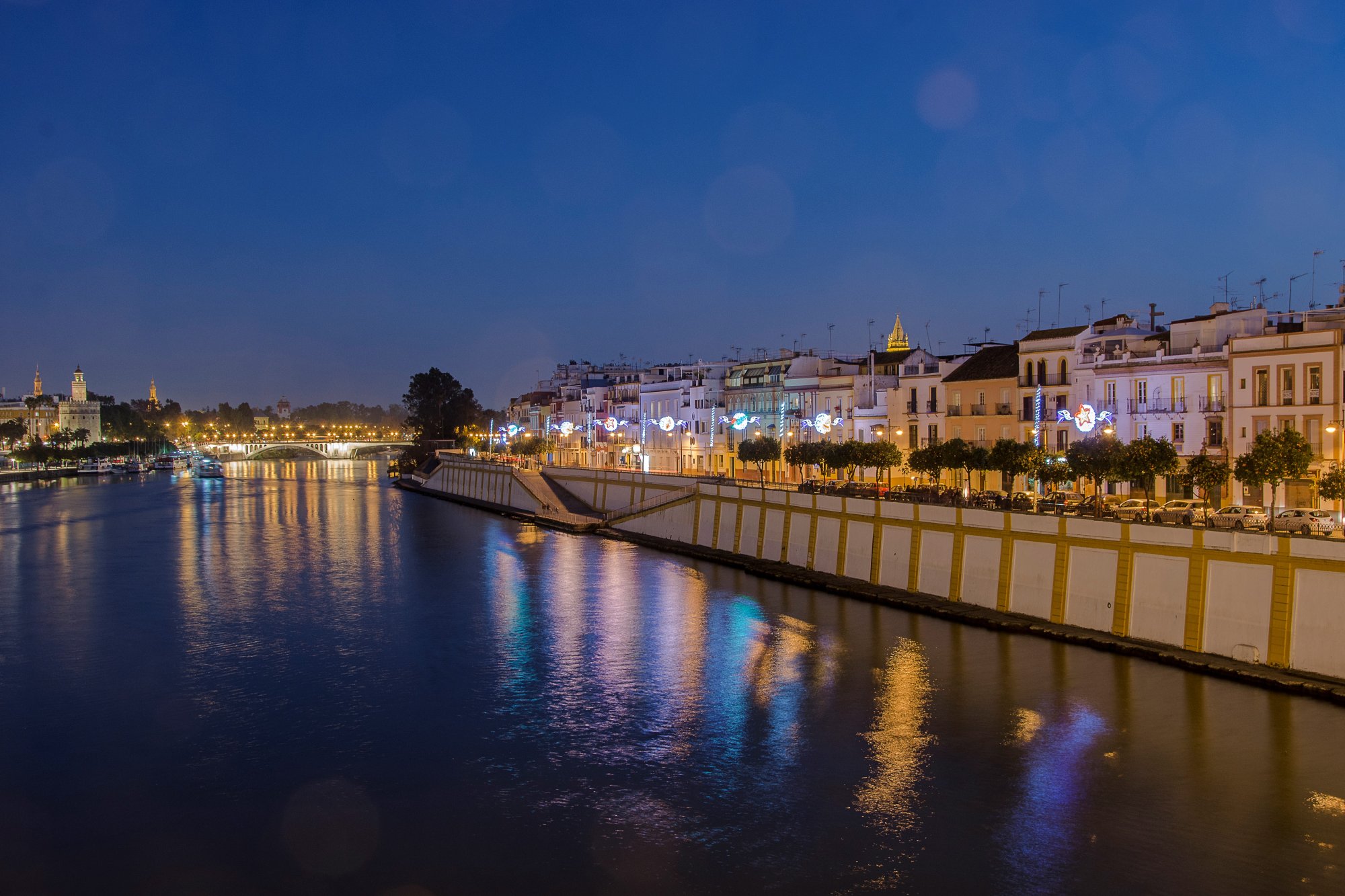 Calle Betis on the Triana riverbank at night, its lit facades reflected in the Guadalquivir