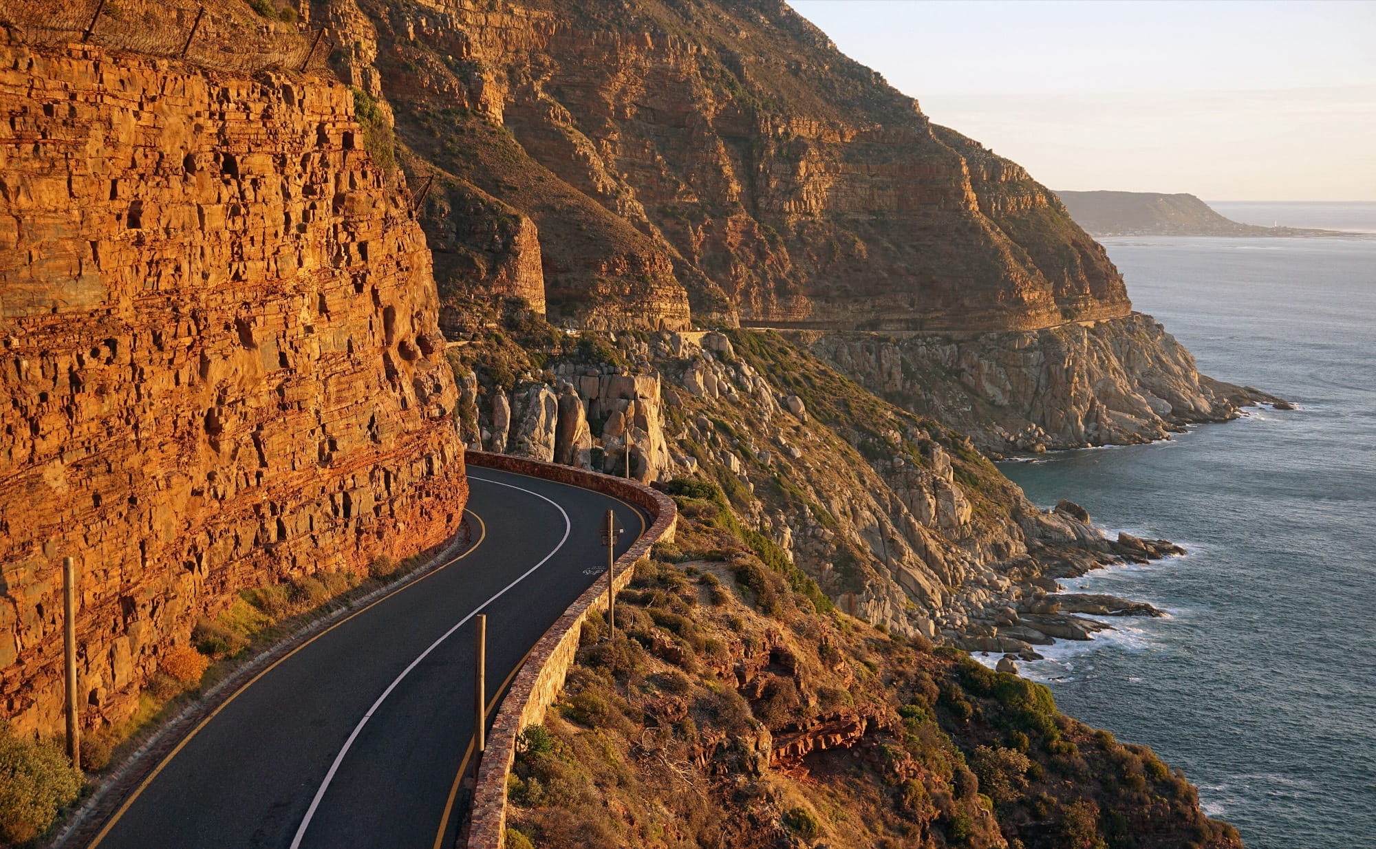 Chapman's Peak Drive curving along a red sandstone cliff above the Atlantic in late-afternoon light, with a low stone wall on the seaward side