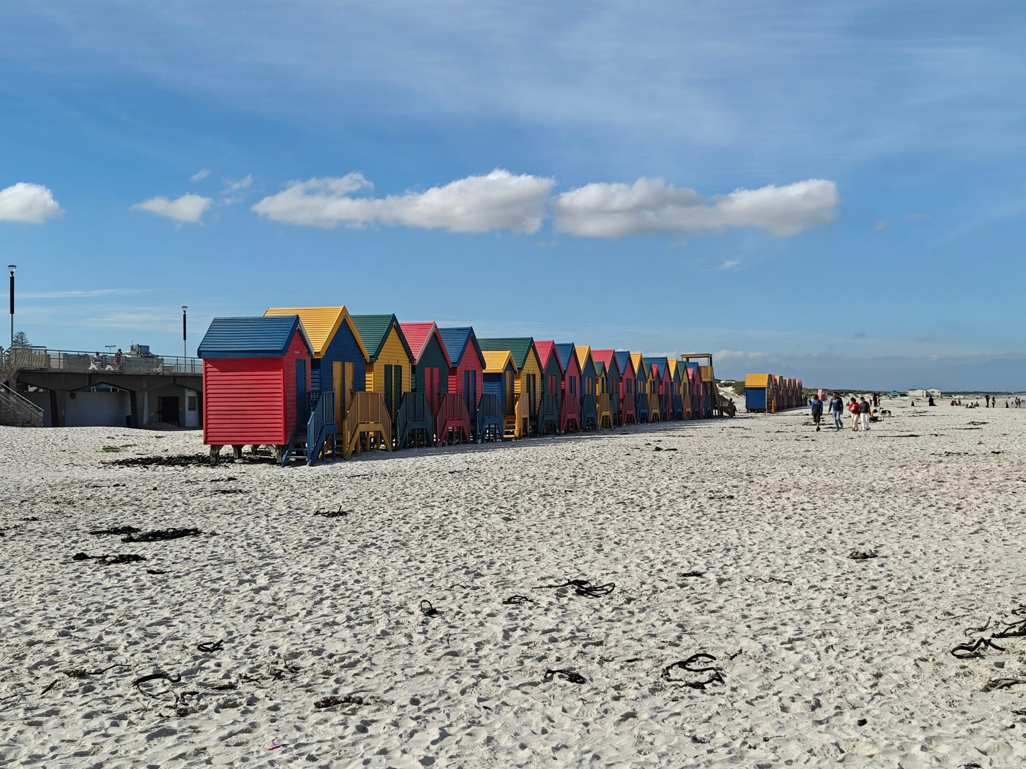 A row of red, yellow, blue and green wooden bathing huts on the wide white sand of Muizenberg beach under a blue sky