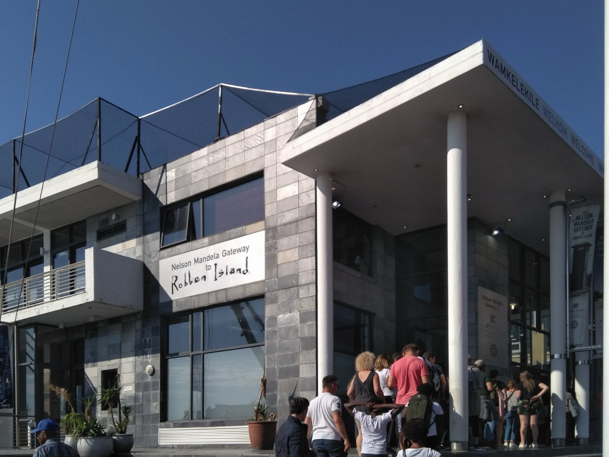 The grey stone facade of the Nelson Mandela Gateway to Robben Island at the Waterfront, with visitors queueing at the entrance under a clear sky