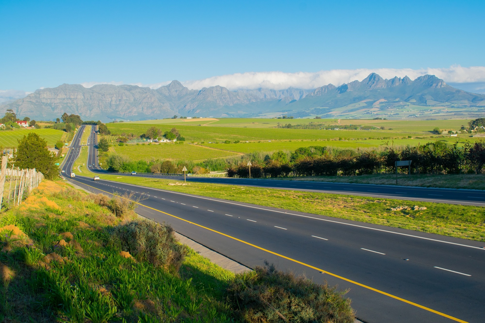 A straight dual carriageway running towards vineyards and the jagged Winelands mountains under a clear sky, on the road from Cape Town to Stellenbosch