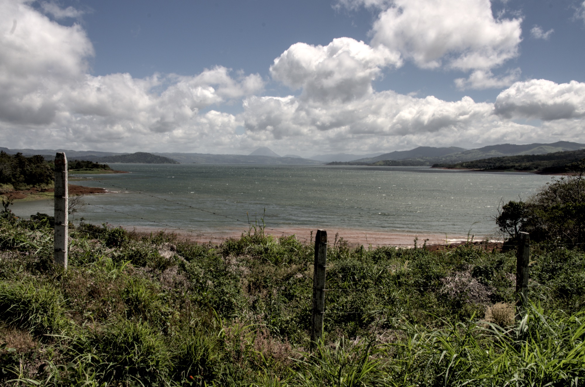 Wind-ruffled grey-green water of Lake Arenal under a cloudy sky, seen over a wire fence and tall grass, with the volcano cone faint on the far horizon