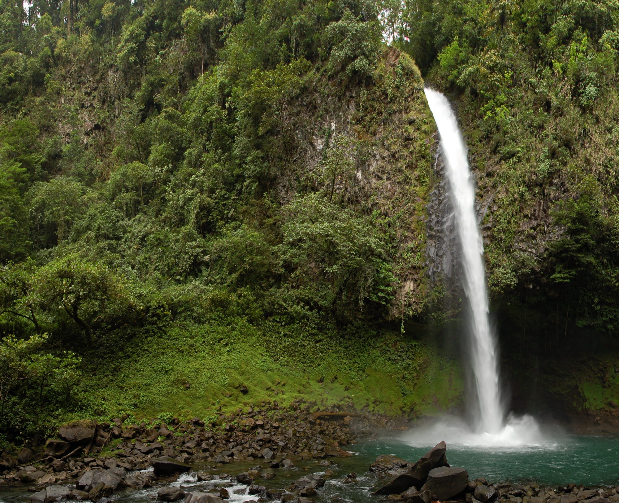 La Fortuna Waterfall dropping in a single white column down a mossy cliff into a turquoise pool with dark boulders in the foreground