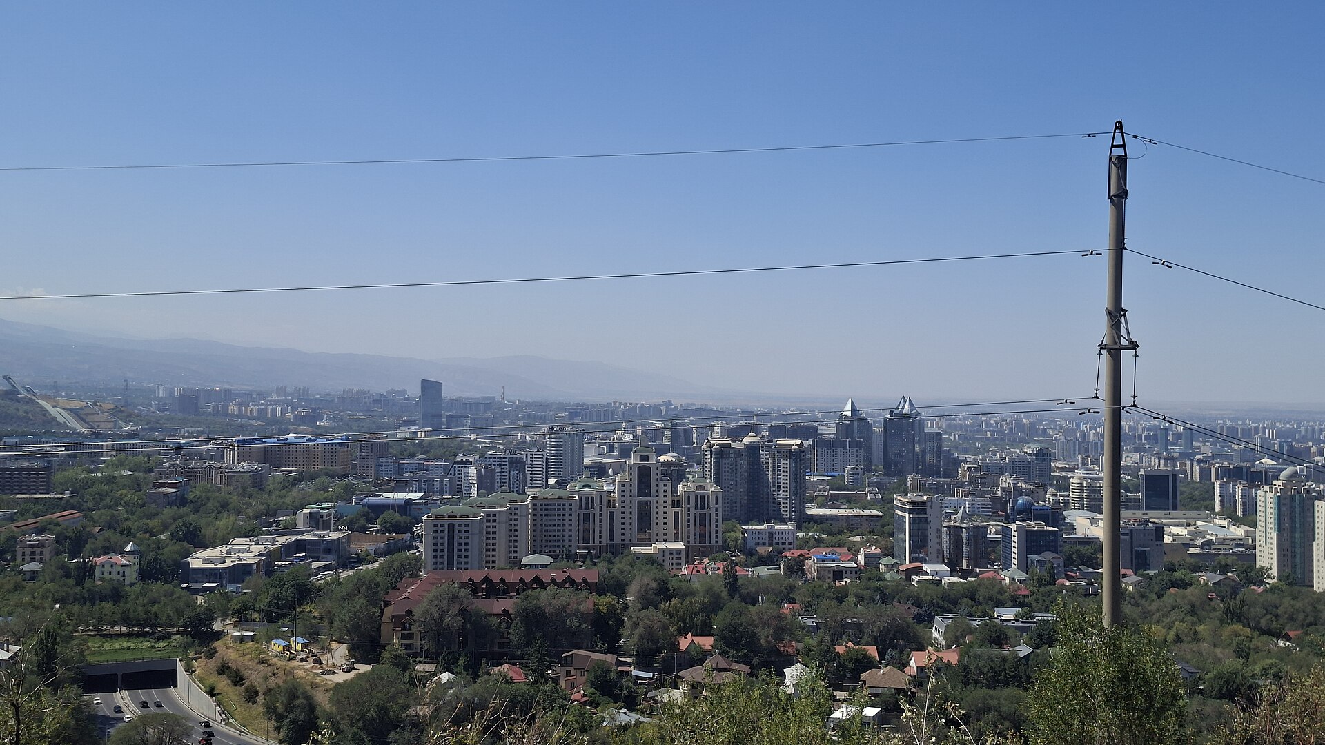 Almaty buildings and tree-lined streets seen from a hillside, with cable lines across the foreground