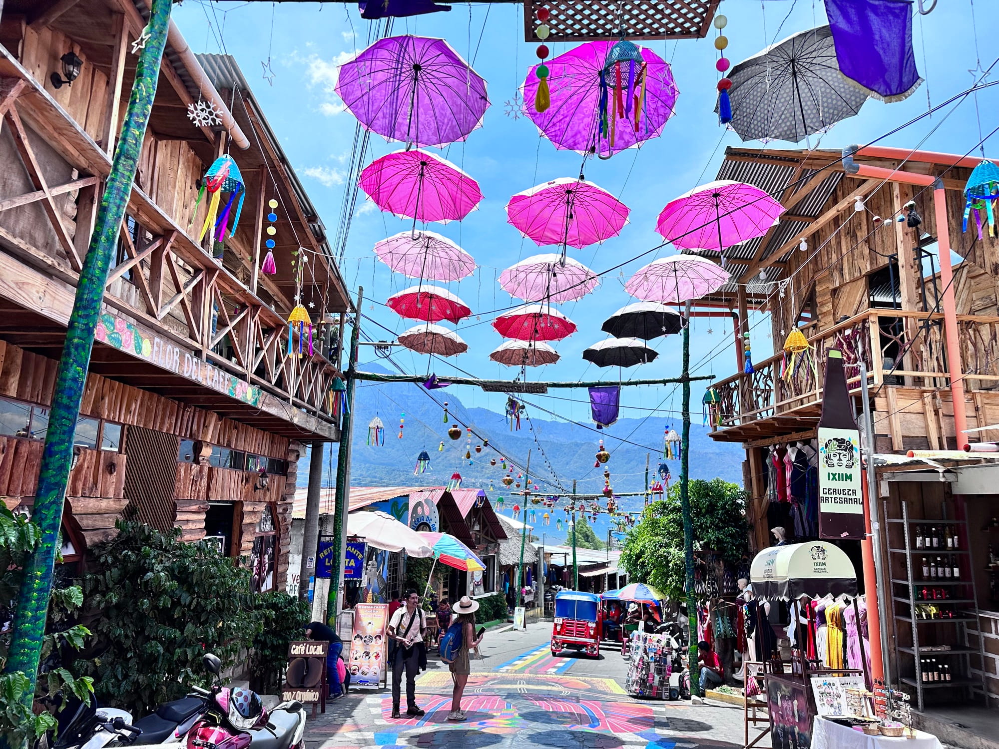 A street in San Juan La Laguna roofed with rows of pink and purple umbrellas between wooden shopfronts, with the lake and hills at the far end