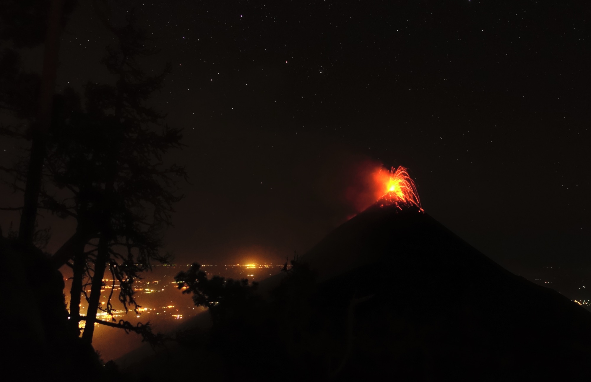 A night-time eruption of Volcan de Fuego throwing red lava from its summit, seen from the Acatenango camp with town lights below