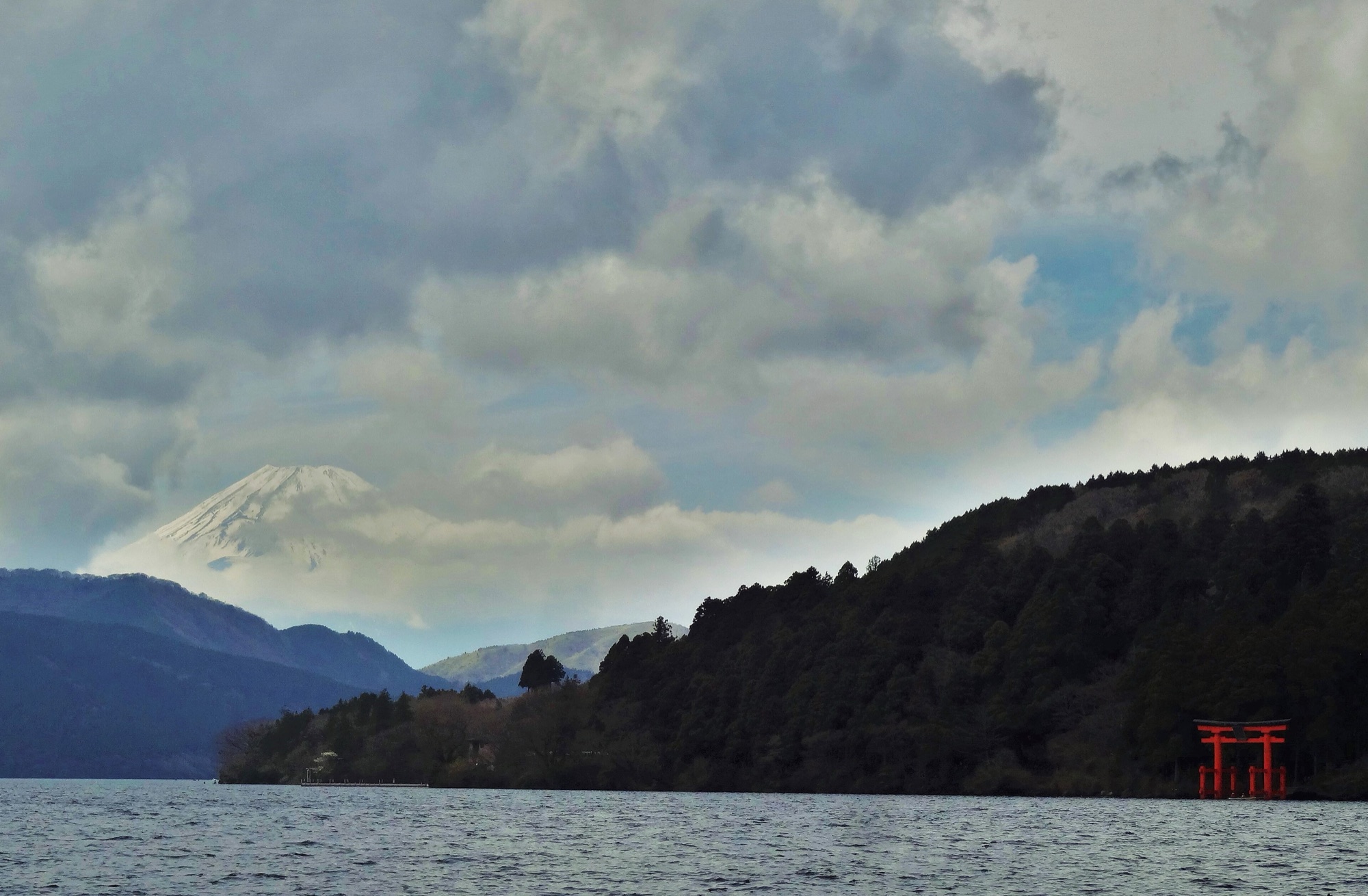 Lake Ashi under a cloudy sky, with the small red torii of Hakone Shrine at the water's edge on the right and the snow-capped peak of Mount Fuji showing between the clouds behind the forested shore