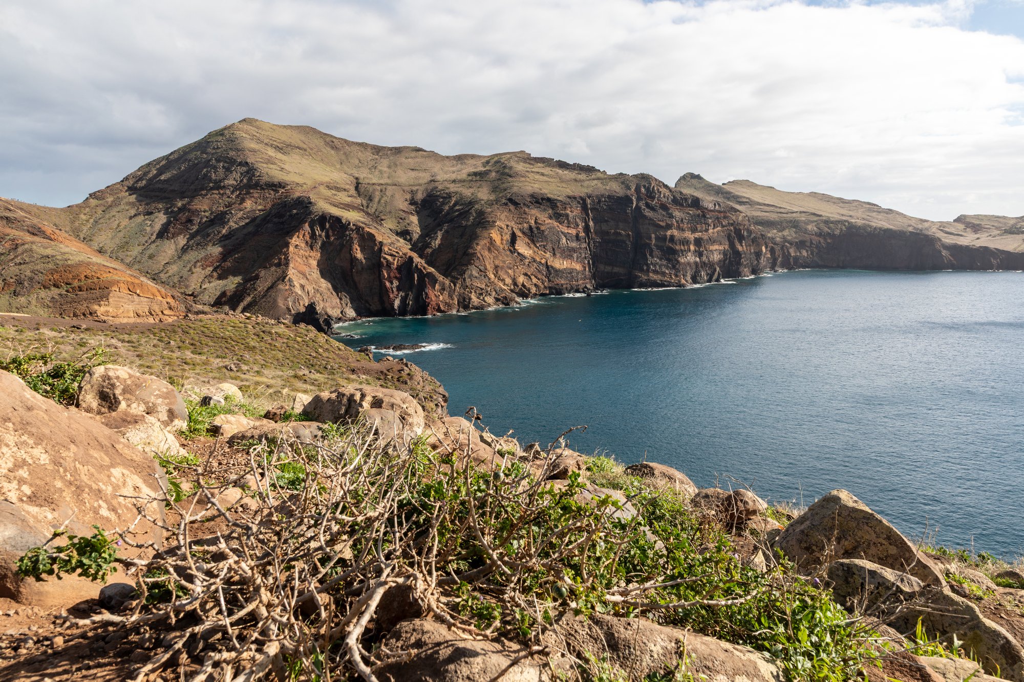 Bare brown cliffs of the Ponta de São Lourenço peninsula above a dark blue bay, with low scrub in the foreground and an overcast sky