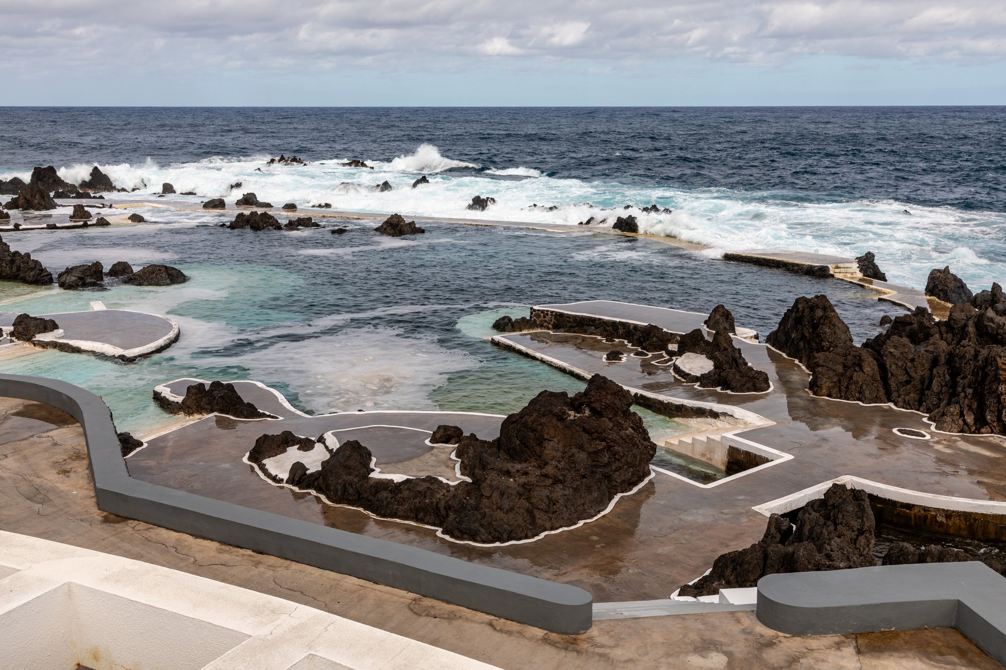 Concrete walkways and lava-rock walls of the Porto Moniz natural sea pools with white surf breaking beyond them