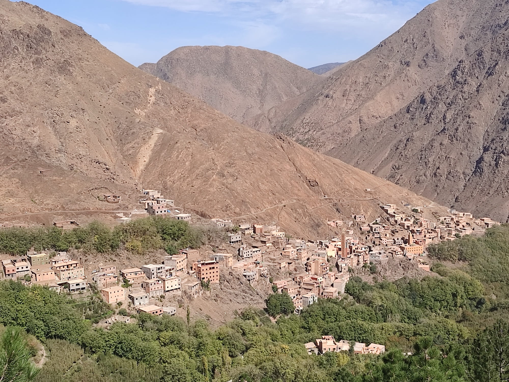 The village of Imlil in the High Atlas seen from above, flat-roofed ochre houses and a square minaret clustered on a green terraced hillside below bare brown mountain slopes under a clear sky