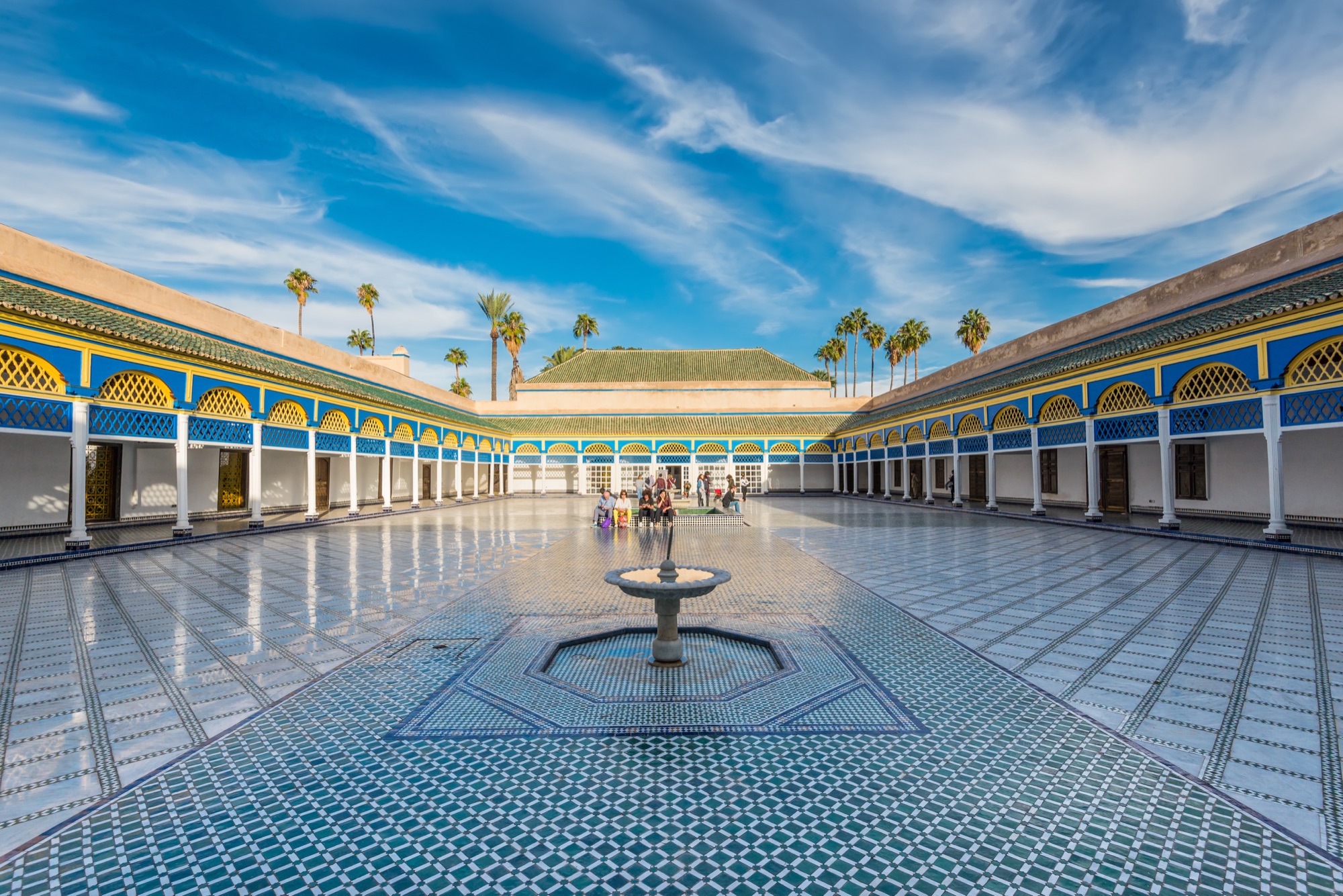 The large open courtyard of the Bahia Palace in Marrakech, paved with green and white zellij tiles around a central marble fountain, with blue, yellow and green painted arcades on three sides and palm trees behind
