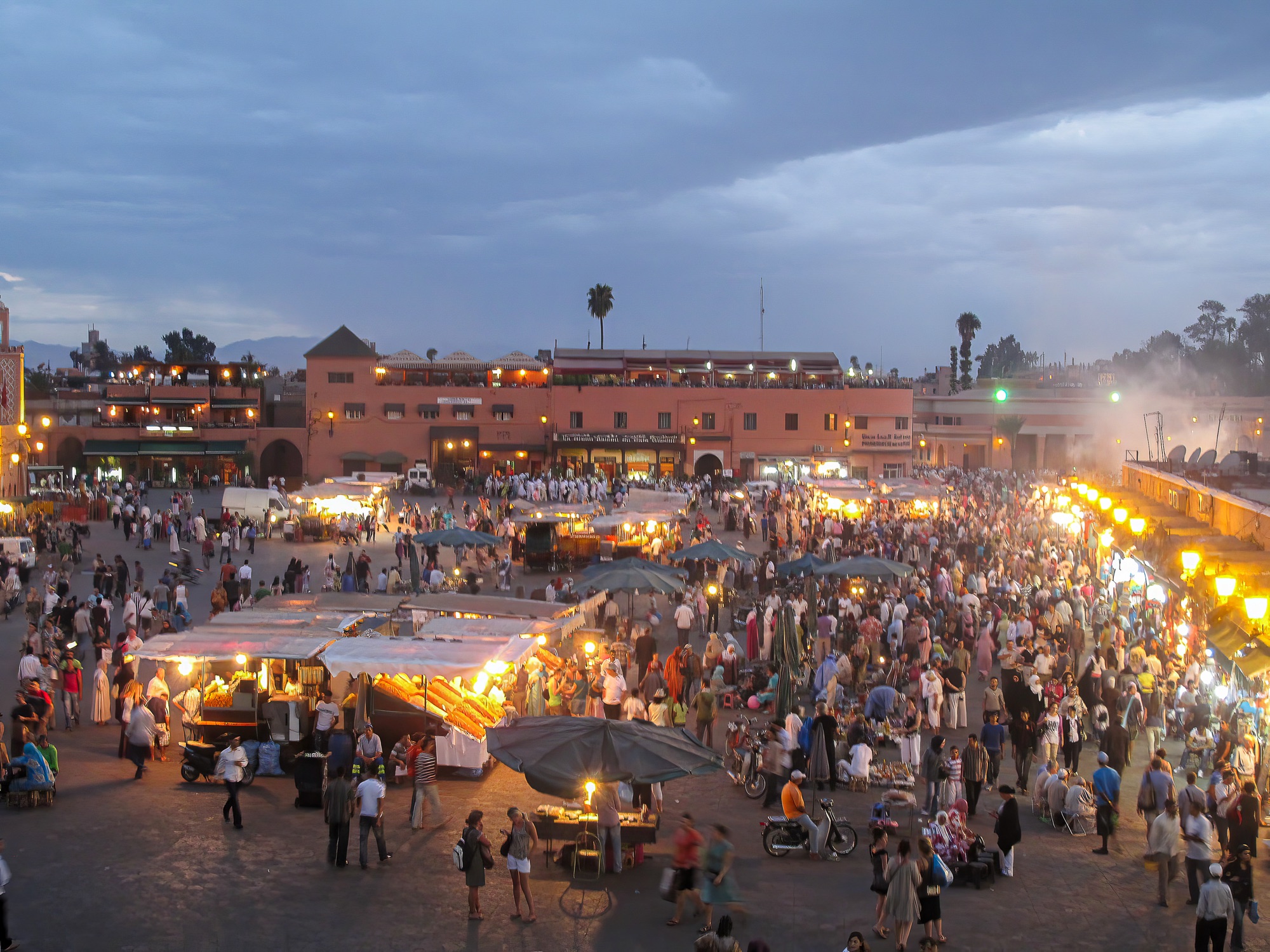Jemaa el-Fna square in Marrakech at dusk, crowds moving between lit food stalls with juice carts, umbrellas and the rooftop terraces of the surrounding cafes under a cloudy evening sky