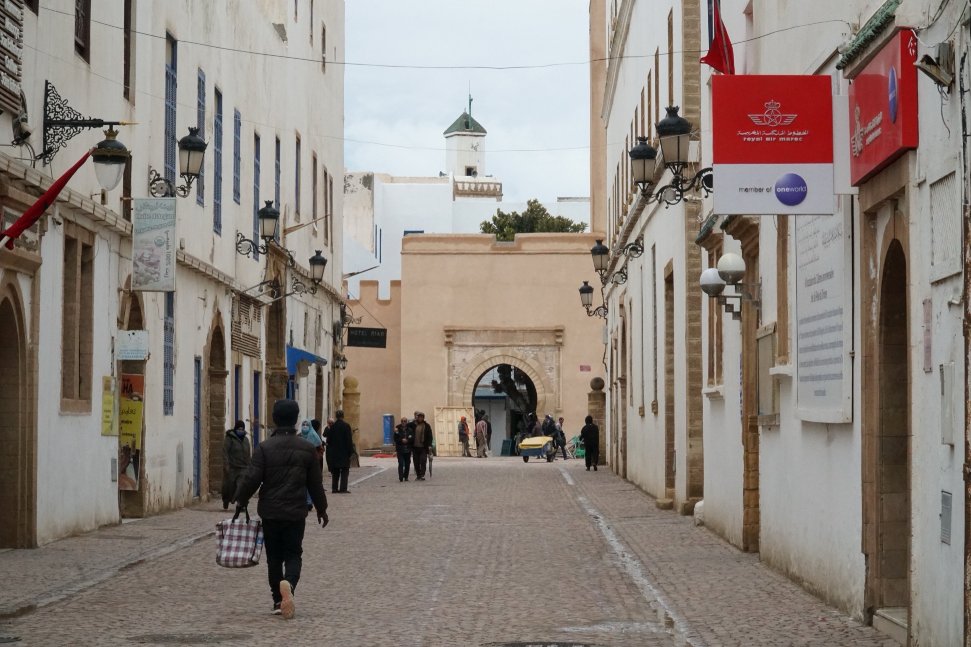 A paved lane in the Essaouira medina lined with whitewashed buildings, blue shutters and wrought-iron lanterns, leading to an arched sandstone gate with a white minaret behind it, a few people walking in winter coats