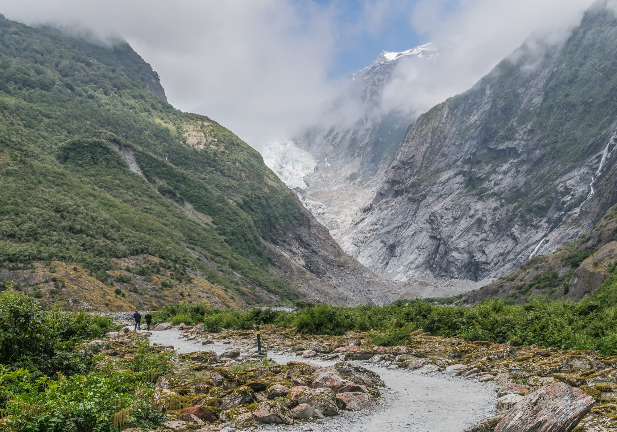 Gravel path through the Franz Josef glacier valley with two walkers heading towards the ice tongue under low cloud between grey rock walls