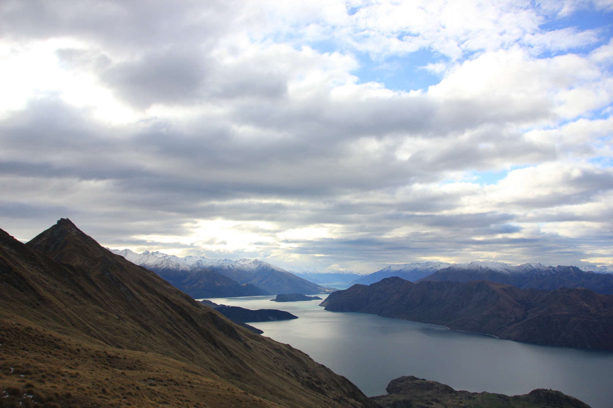 View from the Roys Peak ridge over the golden tussock slopes to Lake Wanaka and snow-capped mountains under a cloudy sky