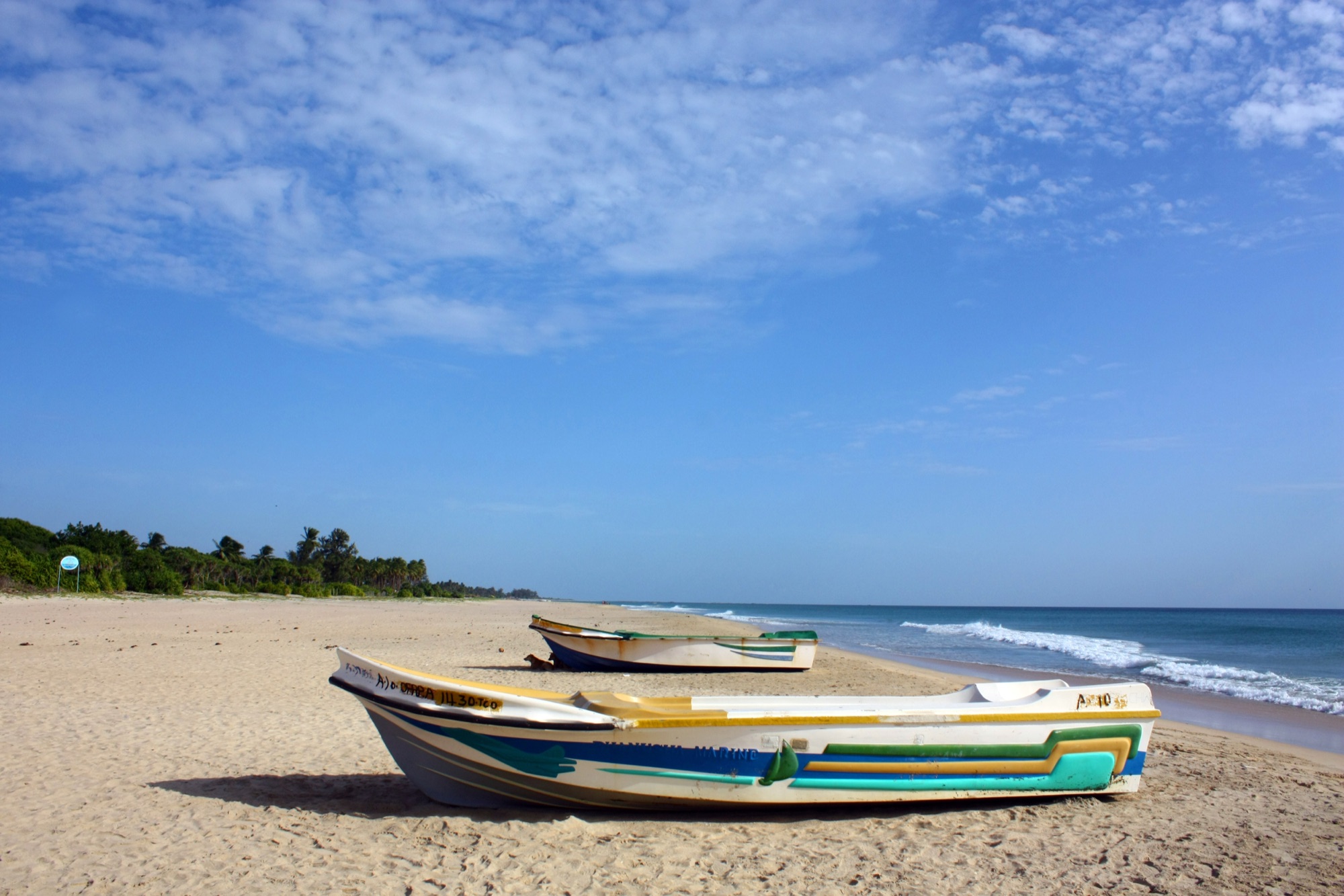 Two painted fishing boats on the wide empty sand of Nilaveli beach near Trincomalee, with small waves and a blue sky