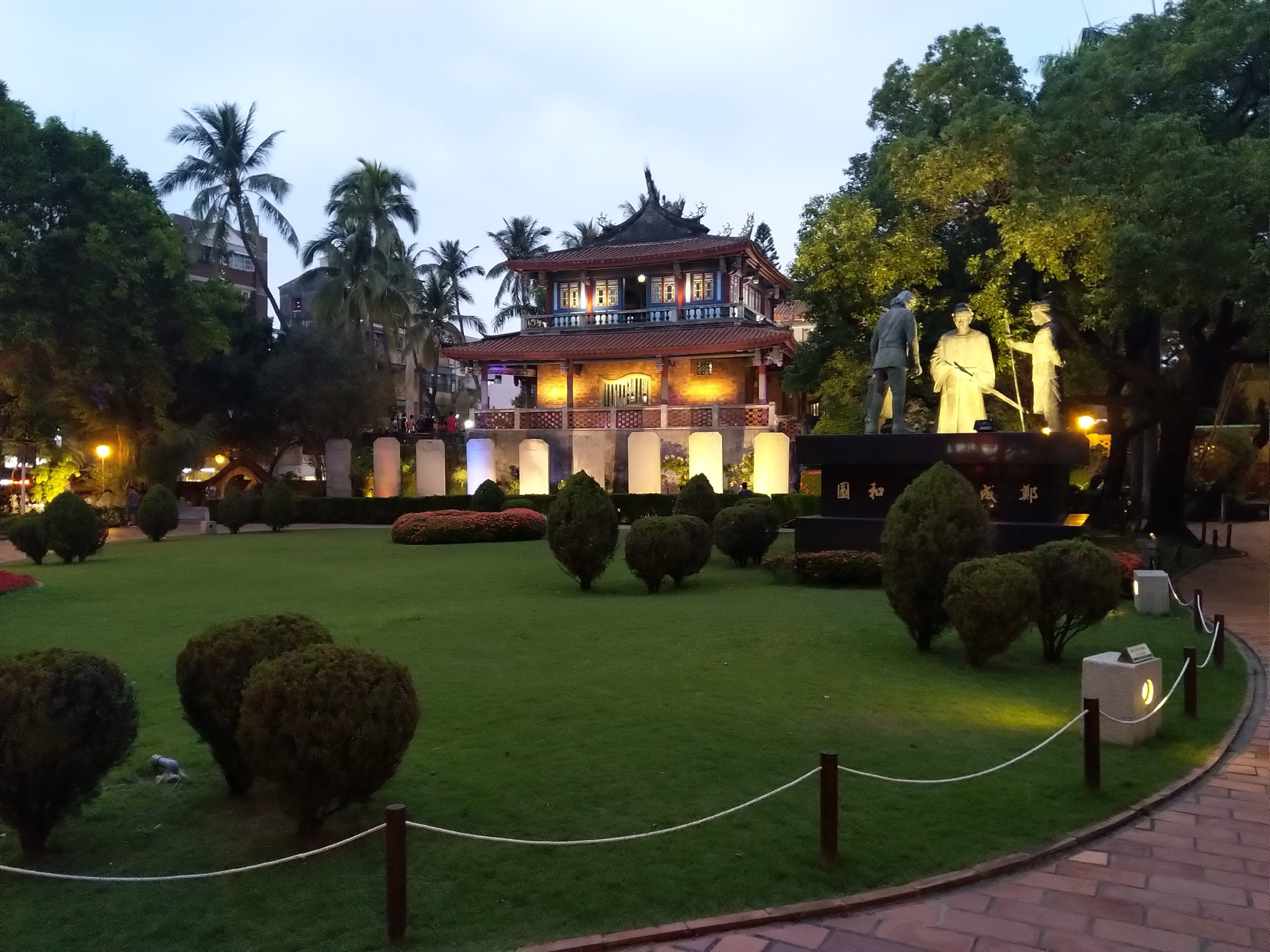 The two-storey red Chihkan Tower lit at dusk behind a lawn with clipped shrubs, palm trees and the statues of the Koxinga negotiation scene