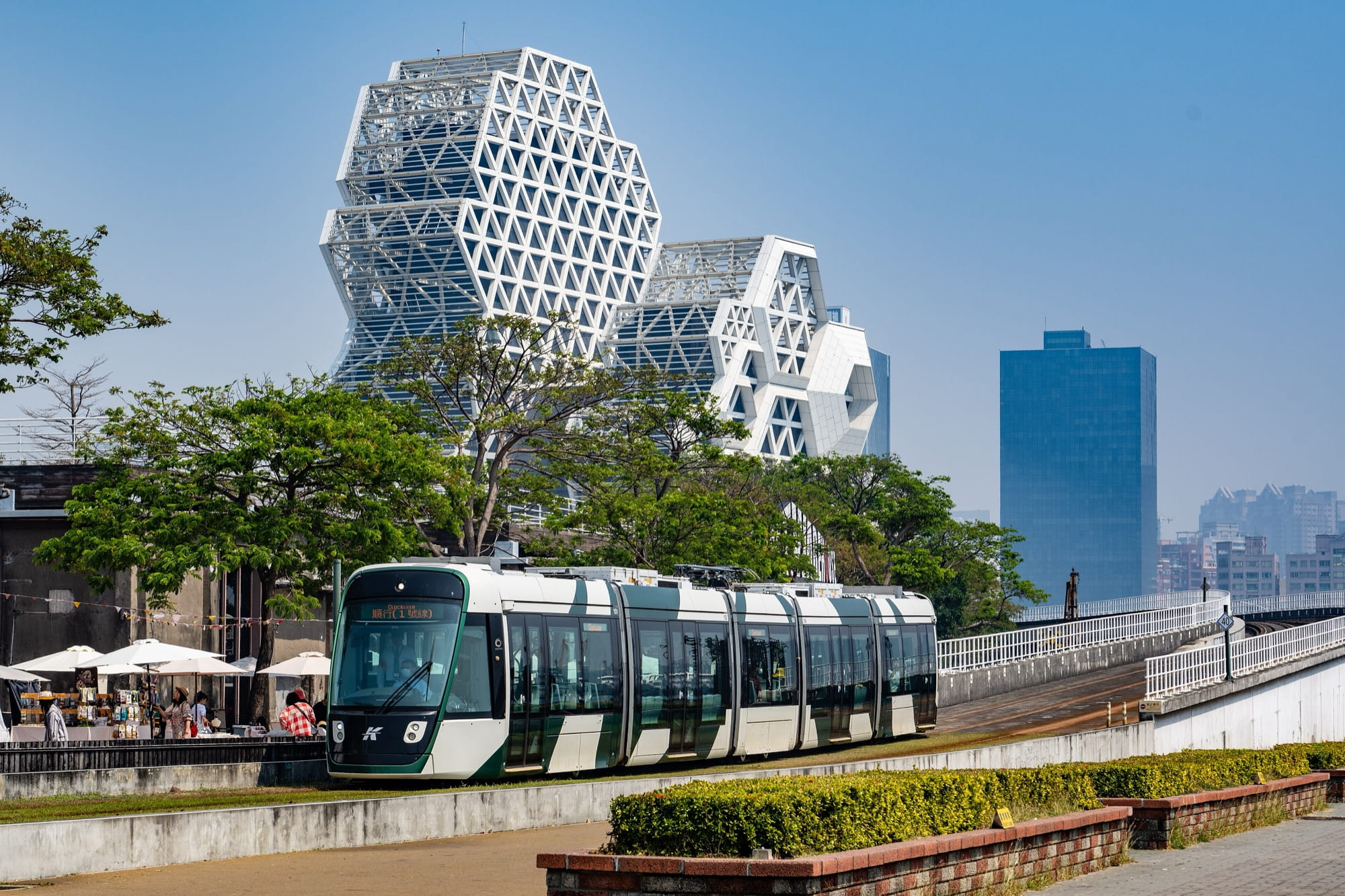A green and white Kaohsiung light rail tram passing the Pier-2 Art Center area with the honeycomb-framed Kaohsiung Music Center behind