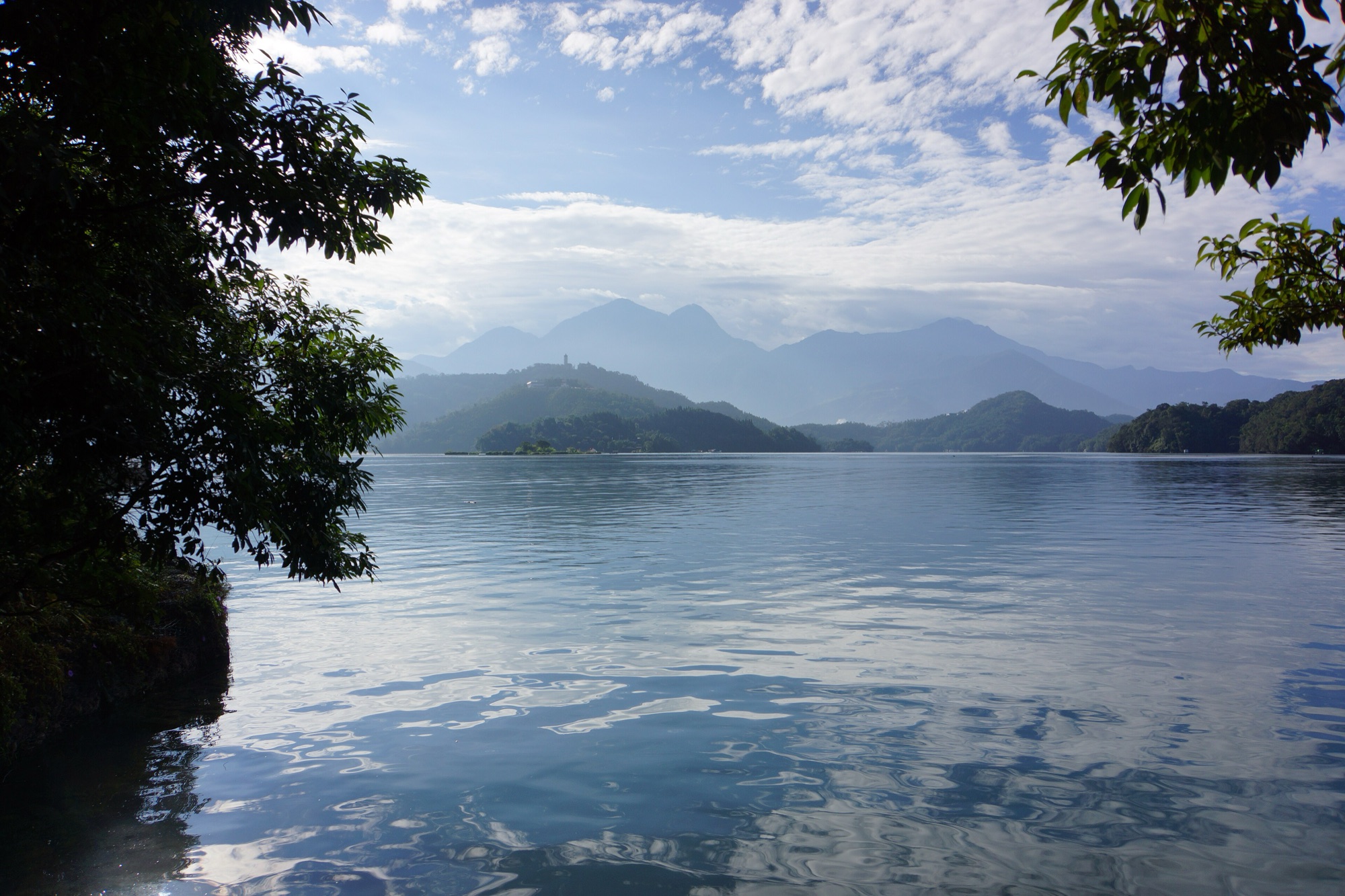 Sun Moon Lake seen from a shaded shoreline path, with Lalu Island and layered blue mountains under a bright hazy sky