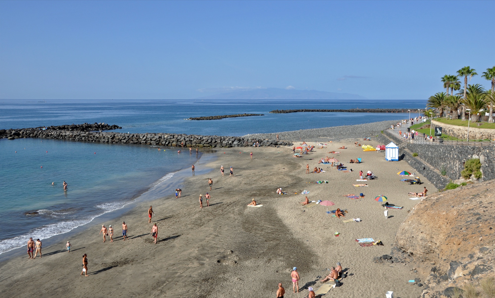 Playa del Duque in Costa Adeje with sunbathers on golden sand, breakwaters in the calm sea and La Gomera on the horizon