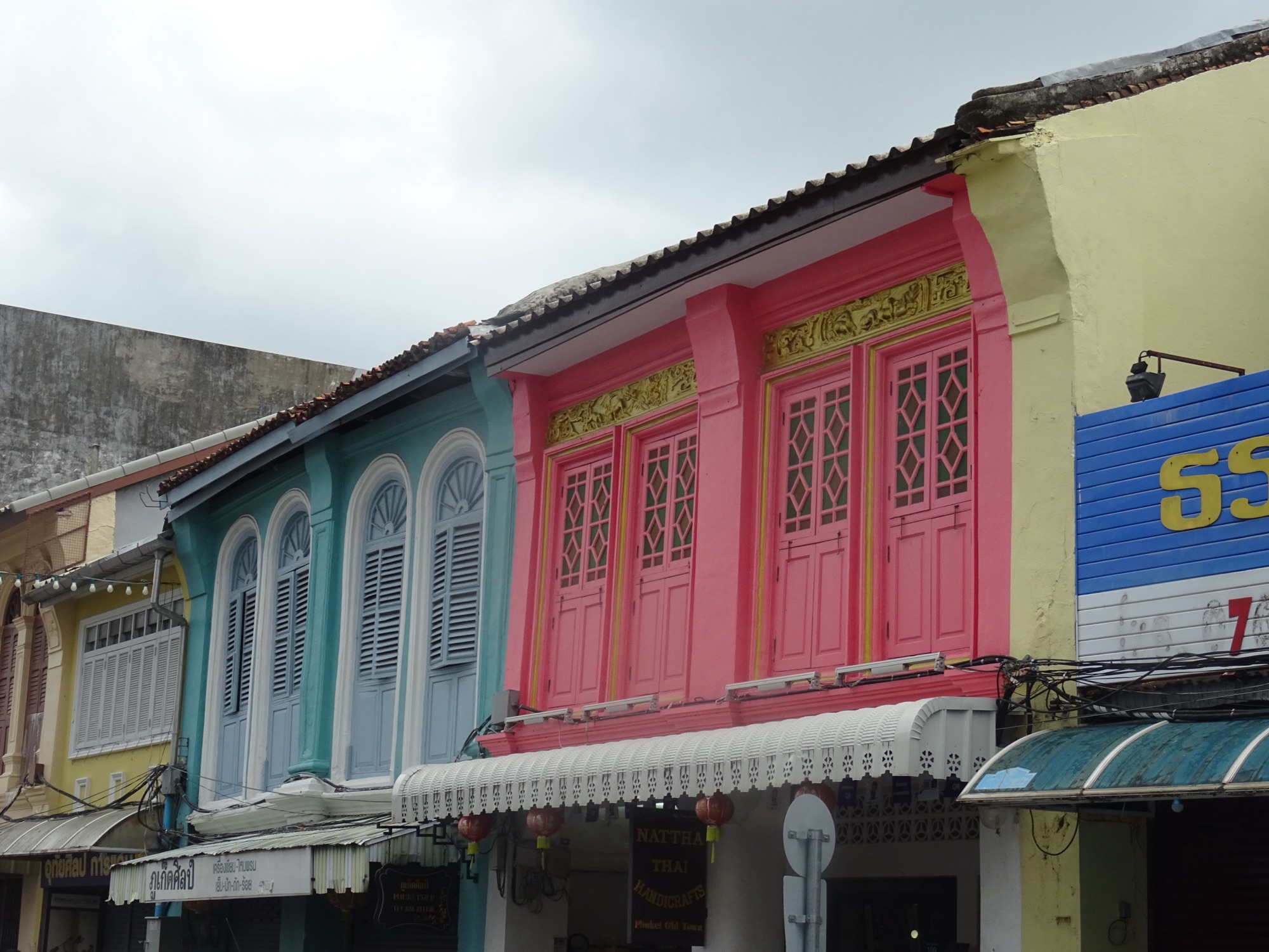 Pastel-painted Sino-Portuguese shophouses on Thalang Road in Old Phuket Town, with a pink facade of arched doors and a blue-grey facade with louvred shutters under an overcast sky
