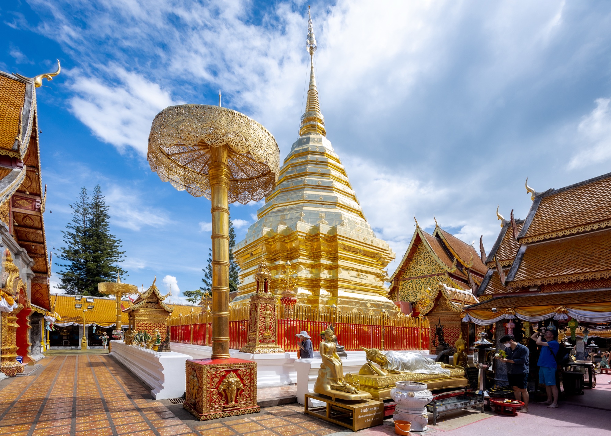 The gilded chedi of Wat Phra That Doi Suthep with a gold ceremonial umbrella and red-and-gold railings under a blue sky with scattered clouds; a few visitors stand near the shrines on the right