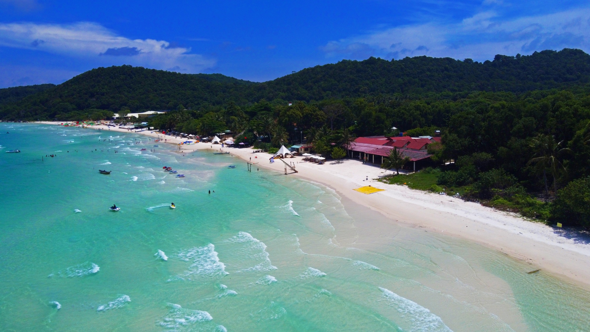 Aerial view of Sao Beach, Phu Quoc: a curve of white sand with turquoise shallows and small waves, a few swimmers and moored boats, a red-roofed beach restaurant and dense green forested hills behind