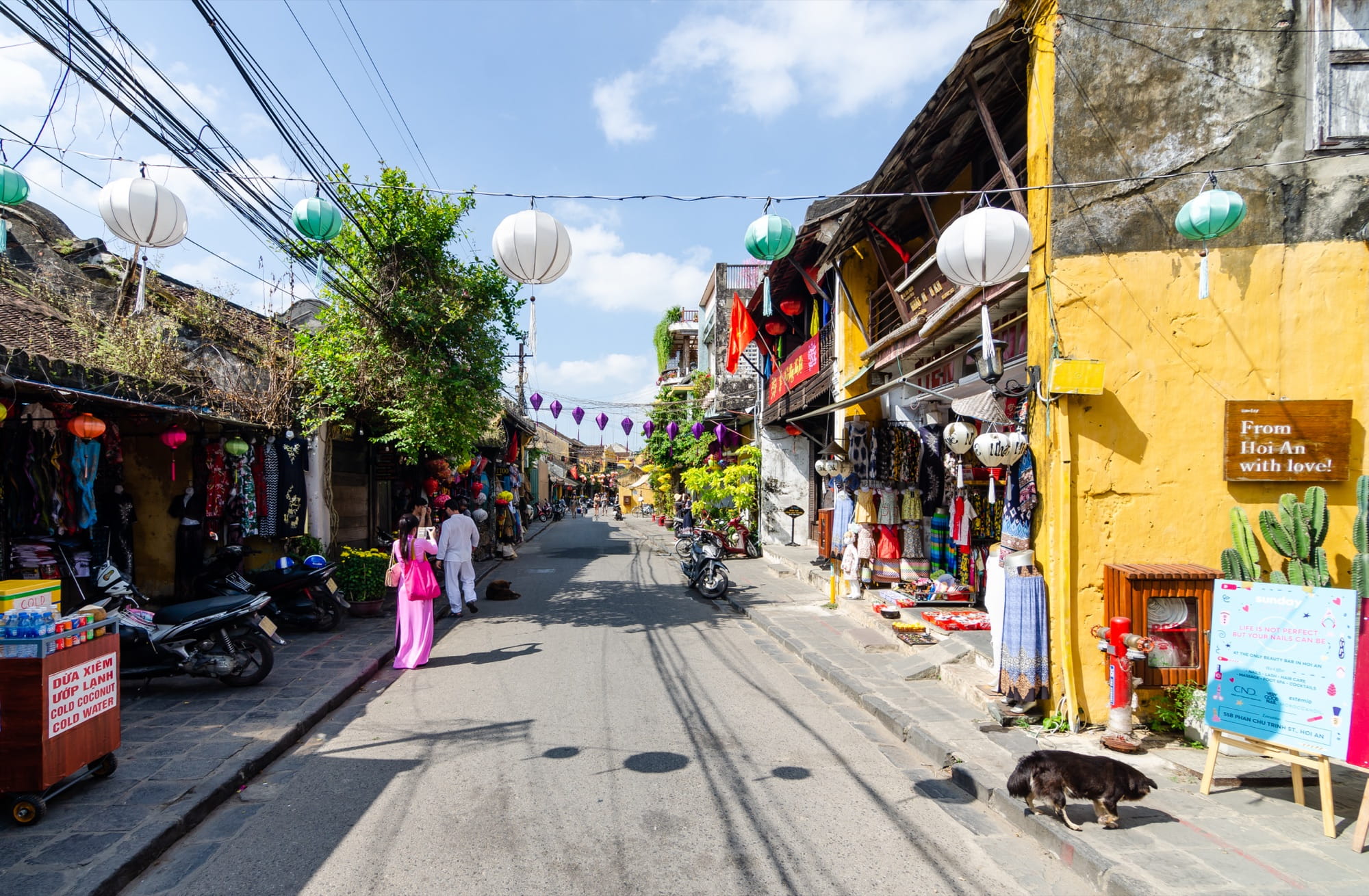 Tran Phu Street in Hoi An Ancient Town under a blue sky: yellow-washed shophouses with tiled roofs, white and green silk lanterns strung across the street, parked motorbikes, souvenir stalls and a woman in a pink ao dai walking away from the camera