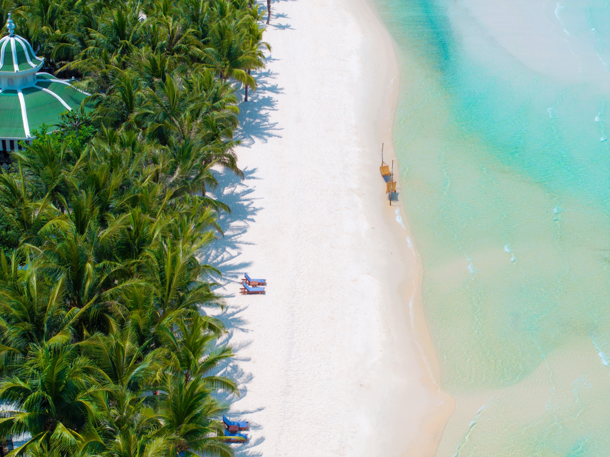 Aerial view straight down onto Kem Beach, Phu Quoc: a strip of white sand between dense coconut palms on the left and pale turquoise water on the right, with a couple of sun loungers, a lifeguard platform and a green-roofed pavilion