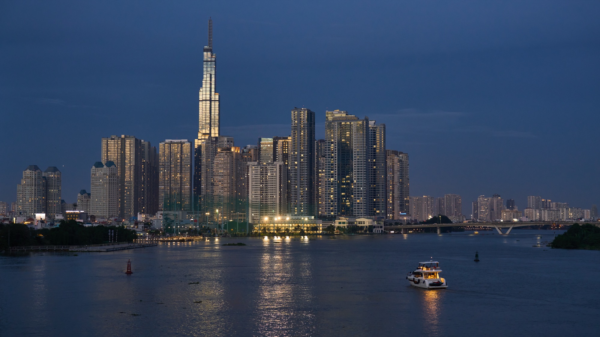 Ho Chi Minh City skyline at dusk from across the Saigon River: the tall spire of Landmark 81 above a cluster of high-rise towers with lit windows, a bridge on the right and a small lit boat crossing the dark water