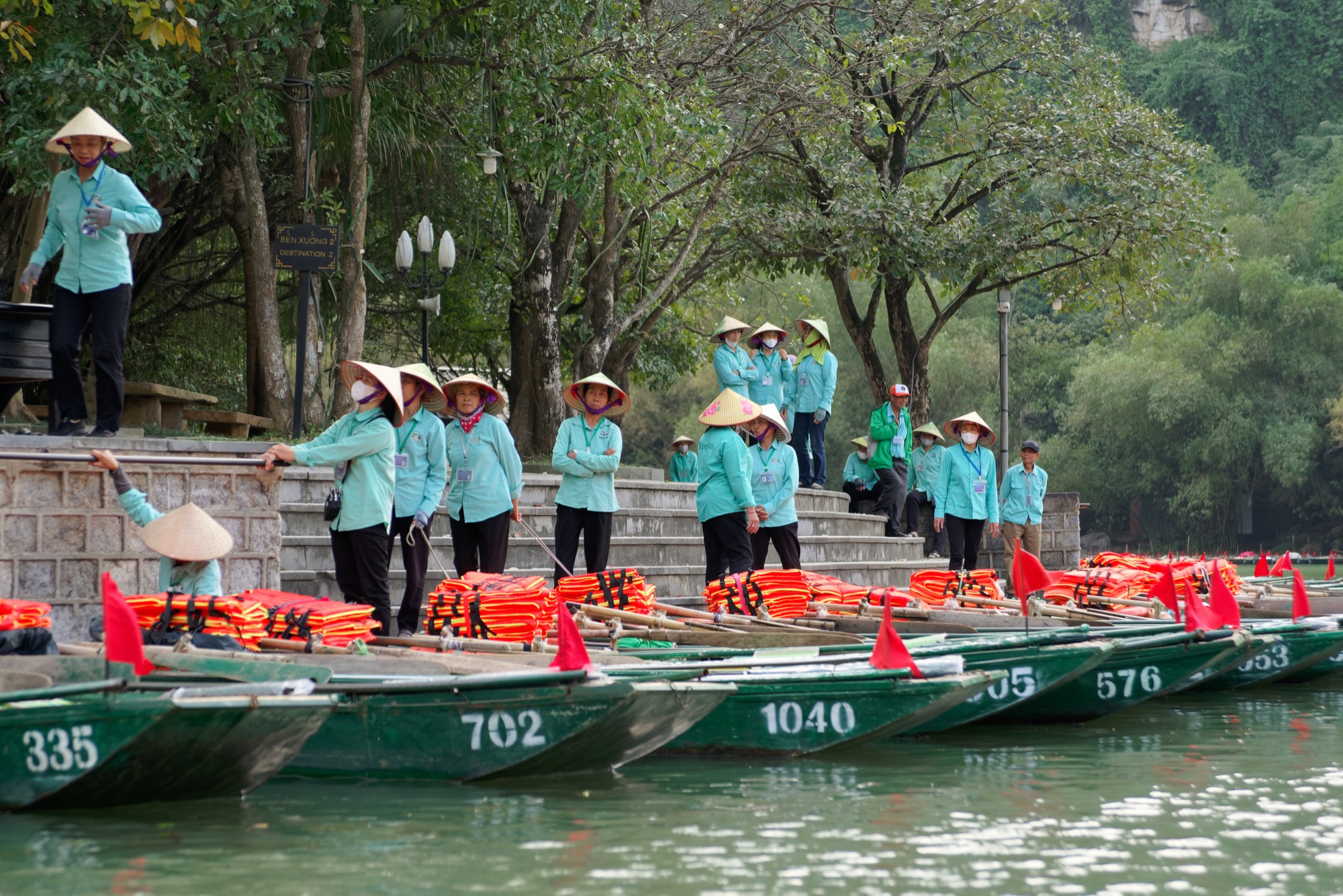 Trang An boat landing in Ninh Binh: rows of green rowing boats with painted numbers and orange life jackets moored at stone steps, a line of rowers in light green uniforms and conical hats waiting on the bank under trees