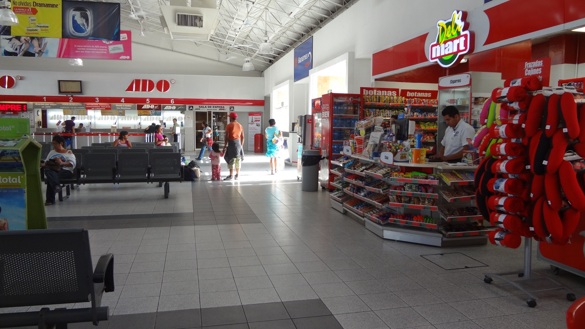 The waiting hall of the ADO bus terminal in Mérida: numbered ticket windows under a red ADO sign, rows of metal seats with a few passengers, and a small shop kiosk on the right