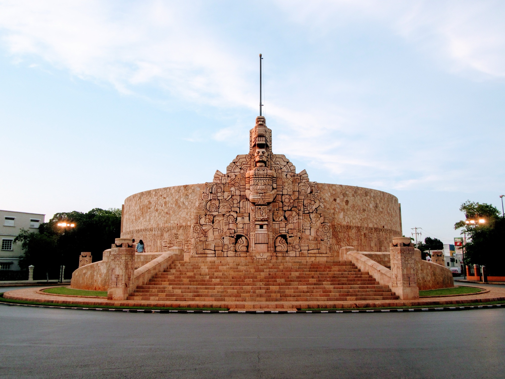Monumento a la Patria on Paseo de Montejo in Mérida: a semicircular monument of carved pink stone with a central Maya-style face and stepped staircases, photographed from the roundabout at dusk
