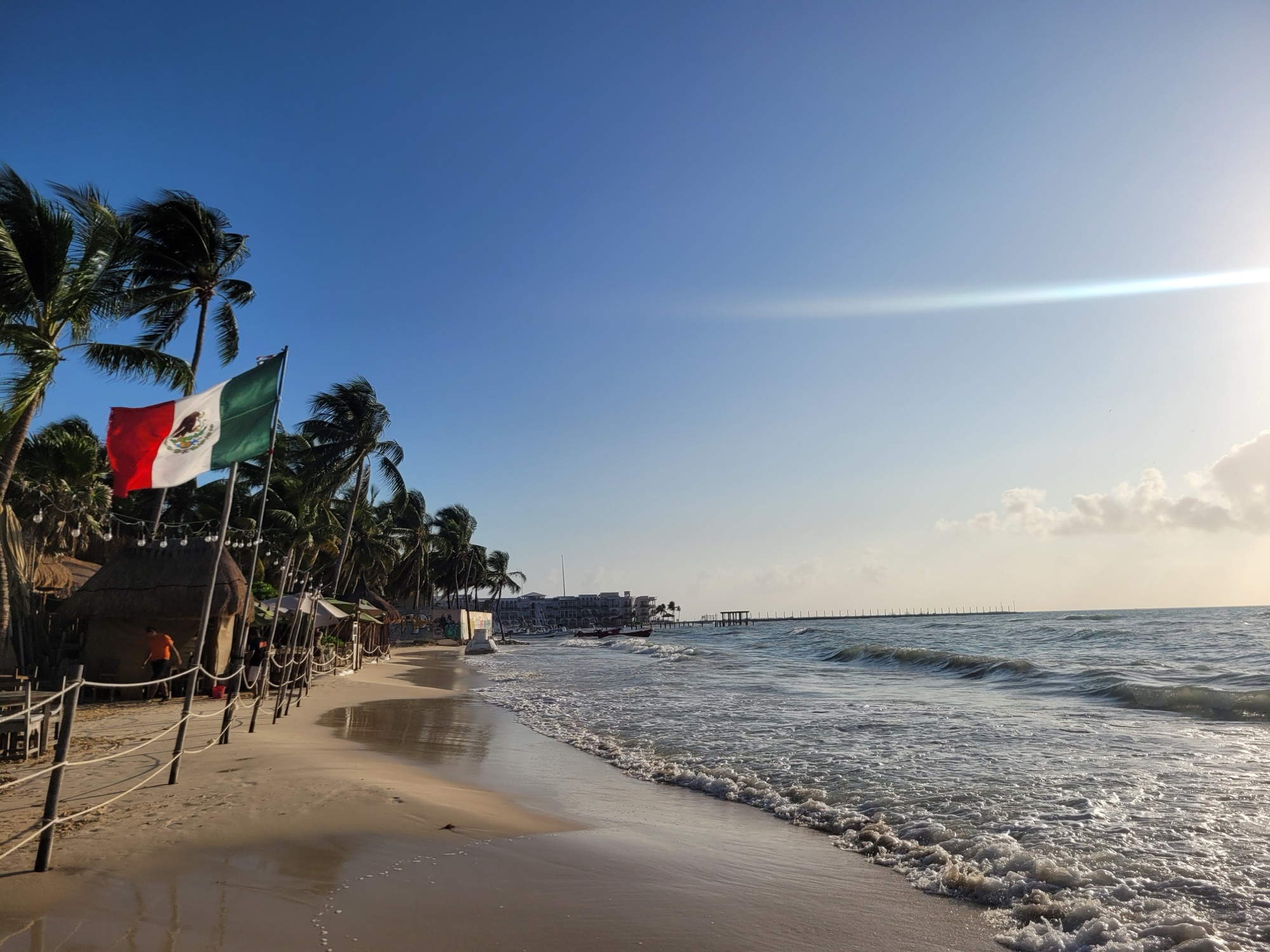 Playa del Carmen beach early in the morning: a Mexican flag and palms on the left, a wide strip of pale sand, small breaking waves and a long pier in the distance under a clear blue sky