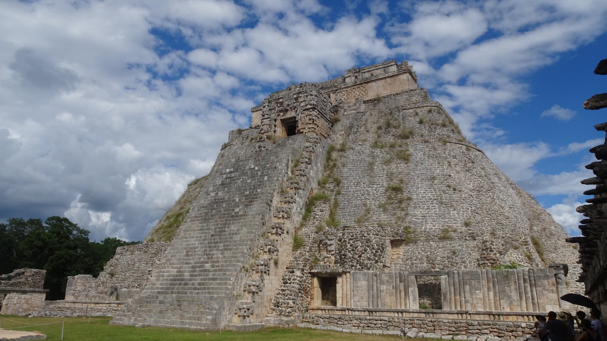 The Pyramid of the Magician at Uxmal: a tall rounded stone pyramid with a steep central staircase and a temple doorway near the top, under a bright blue sky with scattered white clouds