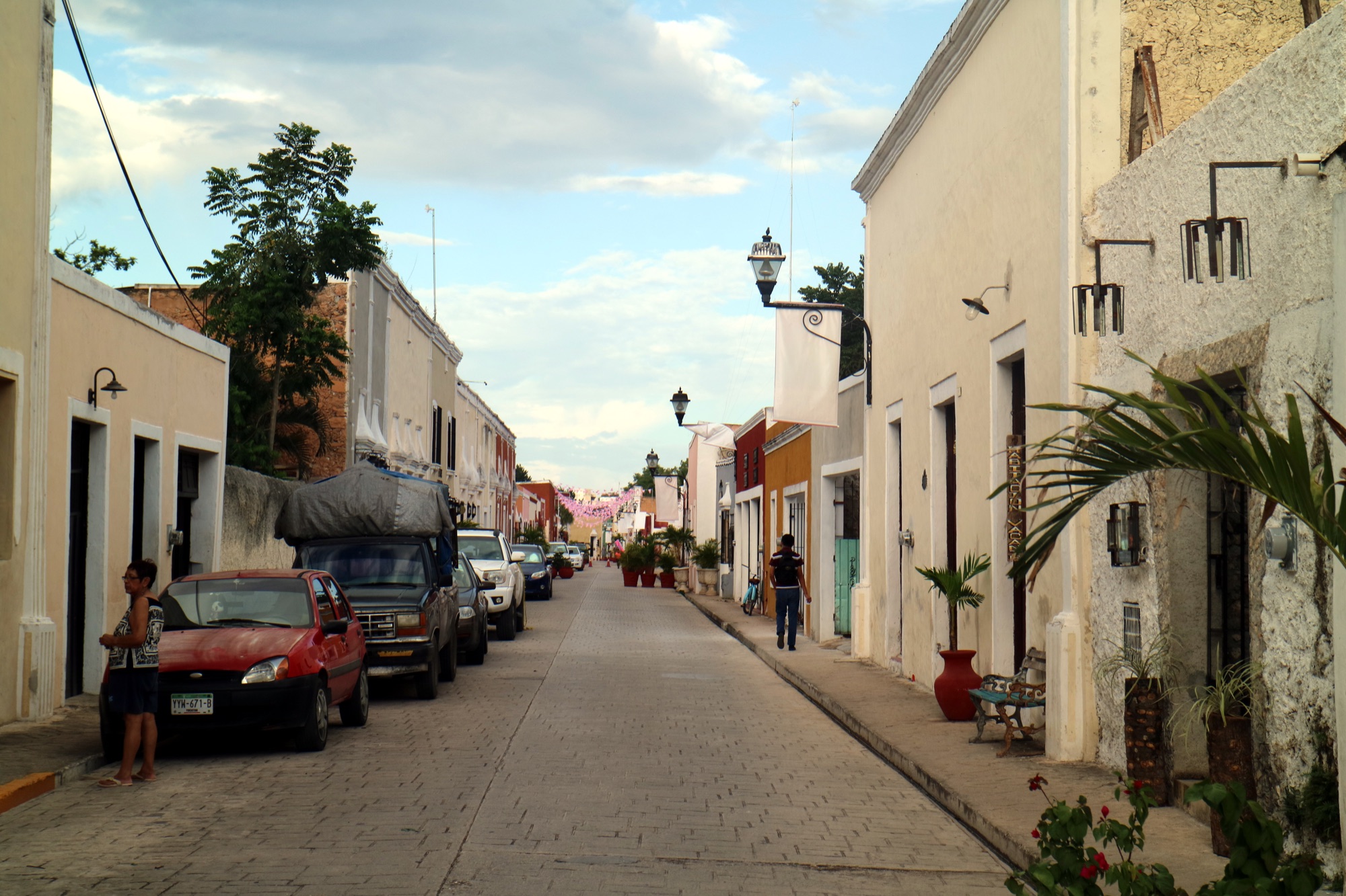 Calzada de los Frailes in Valladolid, Yucatán: a straight cobbled street lined with low cream, yellow and orange colonial houses and iron lanterns, with parked cars on the left and a lone pedestrian ahead